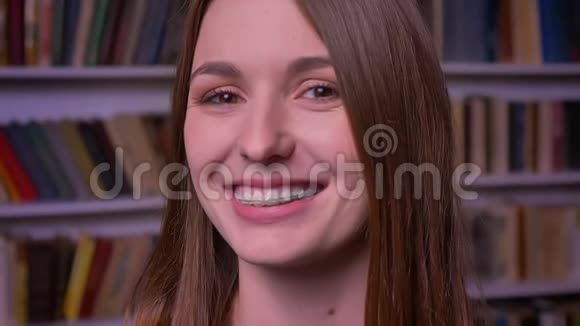 在书架背景上年轻的生姜白种人女孩戴着牙套观看并微笑着进入相机的特写肖像视频的预览图