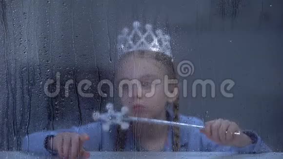 戴着魔法棒的戴着皇冠的小女孩独自坐在雨窗后视频的预览图