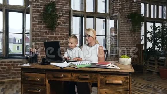 中年漂亮的商务女性坐在木桌旁看着她可爱的孙子玩手提电脑视频的预览图