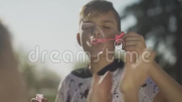 那个穿红色衬衫的男孩在公园里吹着肥皂泡的肖像孩子们的手在泡泡肥皂泡慢动作视频的预览图