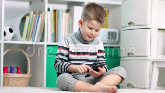 一个小男孩在家坐在地毯上时正在使用他的智能手机视频的预览图