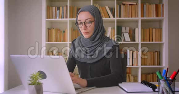 年轻漂亮的穆斯林女商人戴着头巾用手提电脑在室内看着图书馆里的照片视频的预览图