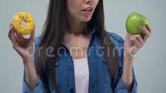 困惑的亚洲女人拿着甜甜圈和绿色苹果健康的食物选择特写视频的预览图