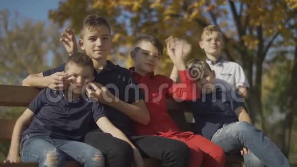 五个男孩坐在长凳上在相机上挥动手朋友们花时间在户外的大公司里视频的预览图