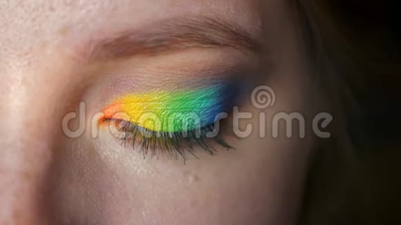 高加索女孩近距离蓝色眼睛上的彩虹妆看着镜头呈现出五颜六色的眼影黑色背景视频的预览图