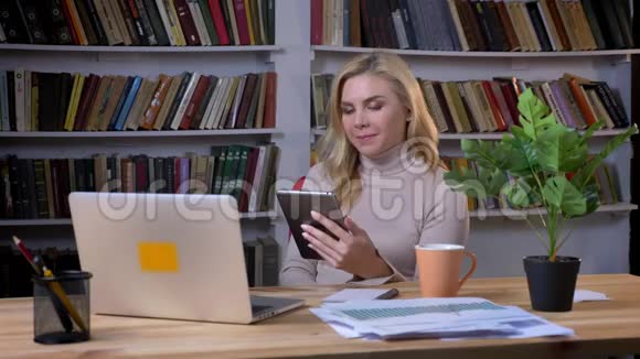 金发中年白种人女商人在书架背景的办公室里与平板电脑合作视频的预览图