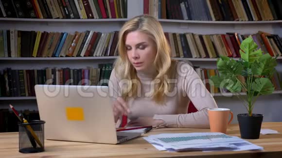 金发中年白种人妇女在书架上的笔记本电脑前窒息和咳嗽的肖像视频的预览图