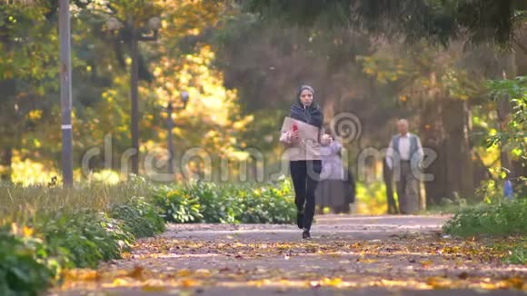 戴着耳机戴头巾的穆斯林妇女与背景人士一起在秋季公园慢跑视频的预览图