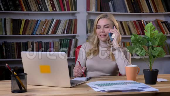 中年白种人金发女郎在手机上说话在书架背景上做笔记的肖像视频的预览图
