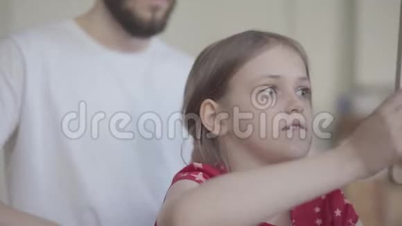 一位留着胡须的年轻父亲正在给他的小女孩梳头而孩子则坐在地上梳理她的玩具视频的预览图