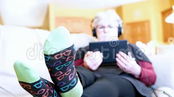 老年妇女在家里的沙发上用平板电脑和耳机放松祖母阅读和听音乐视频的预览图