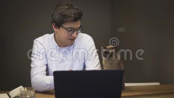 戴着蓝衬衫眼镜的男人坐在桌上拿着笔记本电脑和猫说话视频的预览图