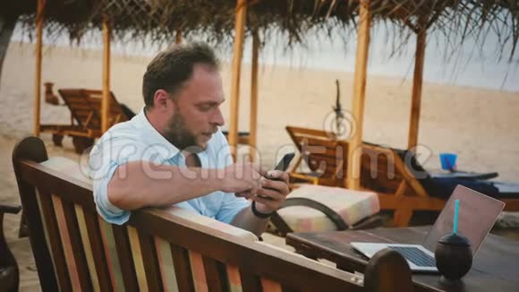 成功的男性自由职业者游客使用智能手机购物应用程序同时坐在夏季海洋度假海滩椅子上视频的预览图
