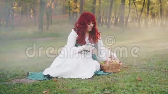 一个神秘孤独的红发女孩坐在地上绿草如茵拿出两个多汁的苹果视频的预览图