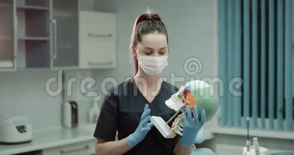 一位年轻的女医生戴着外科口罩和手套在现代诊所的房间里举着一个巨大的人类头骨和视频的预览图