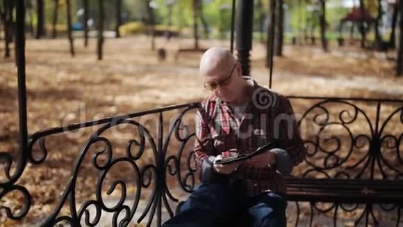 一位戴眼镜的老人坐在秋公园的长凳上看一本电子书视频的预览图