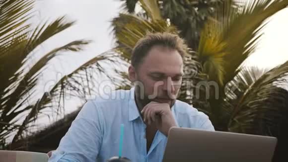 用笔记本电脑和在异国情调的沙滩棕榈树下检查智能手表拍摄快乐微笑的白人商人特写镜头视频的预览图
