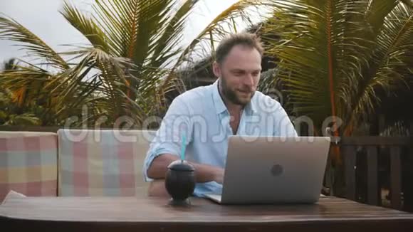 中镜头快乐成功微笑中年商人使用笔记本电脑在异国风情的棕榈树海滩在线工作视频的预览图