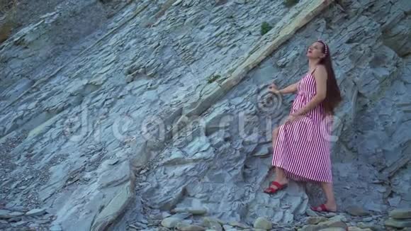 年轻的怀孕女孩穿着条纹红白连衣裙一个女孩站在岩石山脚下抚摸着她的肚子视频的预览图