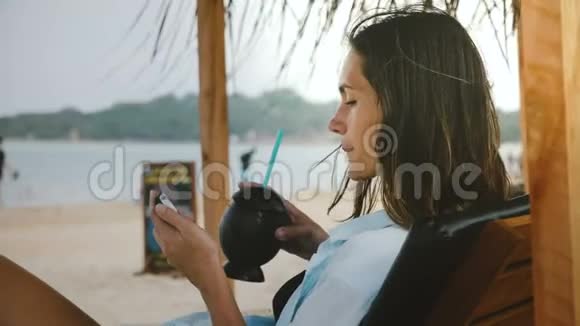 侧景拍摄的年轻放松的自由职业者妇女与异国情调的饮料观看视频智能手机在休闲海滩椅子视频的预览图