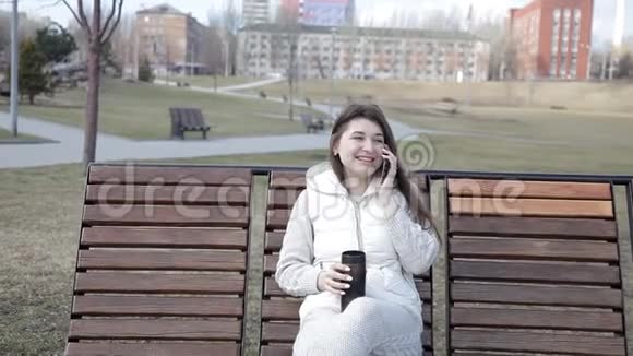 公园里漂亮的女孩坐在长凳上喝着杯子里的咖啡在电话里聊天视频的预览图