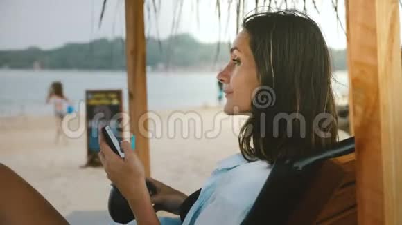 侧面拍摄的年轻快乐微笑的旅游妇女与饮料和智能手机休息环顾海滩椅子视频的预览图