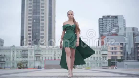 美丽的漂亮女孩穿着一件漂亮的晚绿色连衣裙走在摩天大楼附近的空城广场上真实的人视频的预览图