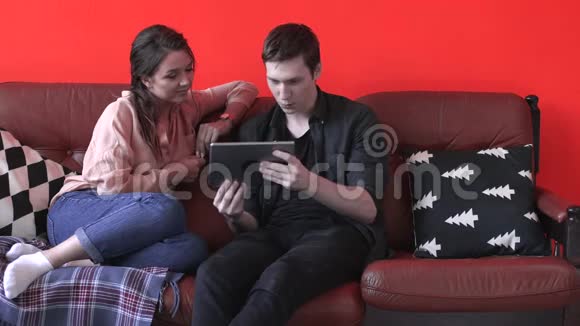 一对幸福的年轻夫妇坐在家里的棕色沙发上看着平板电脑屏幕库存数据视频的预览图
