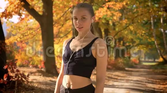 运动的白种人女孩带着耳机在秋季背景上的相机上摆姿势视频的预览图