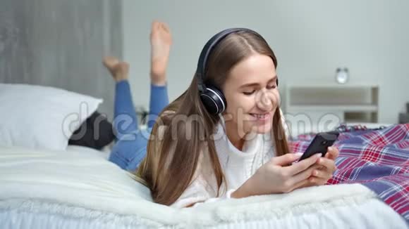 光着脚的年轻漂亮女孩躺在床上用耳机和智能手机听音乐视频的预览图
