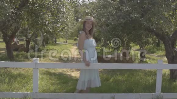 可爱的年轻女子戴着草帽穿着白色长裙看着镜头微笑地站在绿色的夏日花园里视频的预览图
