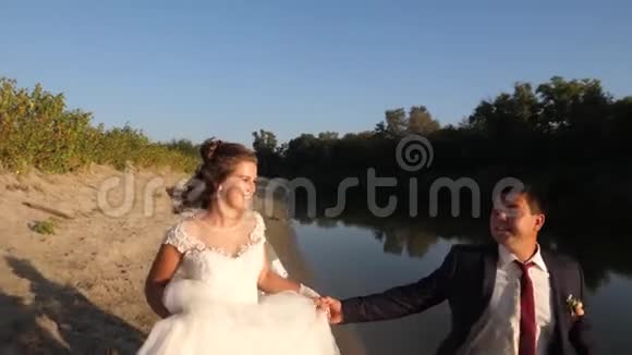 美丽的新娘穿着白色的裙子新郎穿着西装赤脚在河边的海滩上奔跑特写视频的预览图