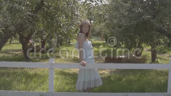 时尚的年轻女子戴着草帽穿着白色长裙站在篱笆后面的绿色夏日花园里农村视频的预览图