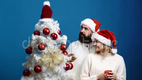 戴着顶帽子的大胡子男人和金发女人用蓝色背景装饰圣诞树可爱的一对准备视频的预览图