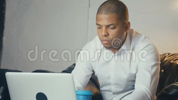 美国黑人商人在现代办公室里坐在沙发上看显示器时使用笔记本电脑和咖啡视频的预览图