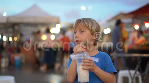 亚洲街头市场上一个小男孩喝椰子奶昔的慢镜头视频的预览图