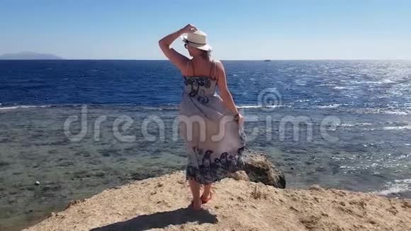 在一个风和日丽的日子里女人站在海边的山上顶着帽子迎风度假视频的预览图