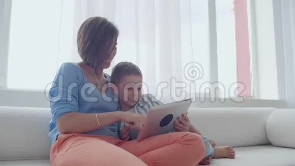 母亲和儿子坐在沙发上使用数字平板电脑快乐的妈妈和小男孩一起使用带有触摸屏的平板电脑视频的预览图