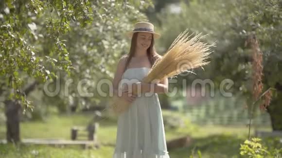 可爱的时尚女青年戴着草帽穿着白色长裙看着镜头微笑地站在绿色的夏天视频的预览图