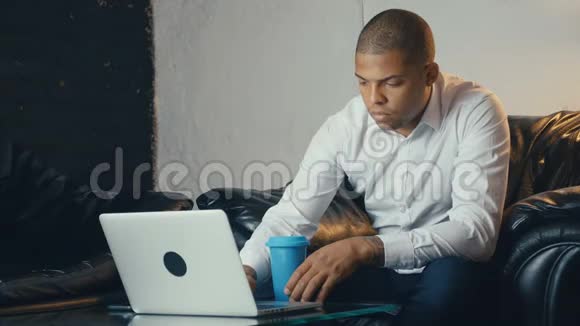 美国黑人商人在现代办公室里坐在沙发上看显示器时使用笔记本电脑和咖啡视频的预览图
