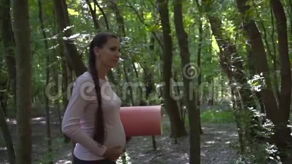 做瑜伽的孕妇一个女孩走在公园里手里拿着地毯另一只手放在肚子上视频的预览图