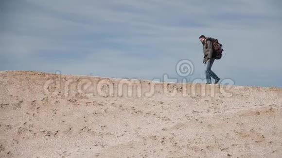 一位戴着眼镜的游客带着一个大背包在美丽的蓝天背景下沿着一个沙丘散步视频的预览图