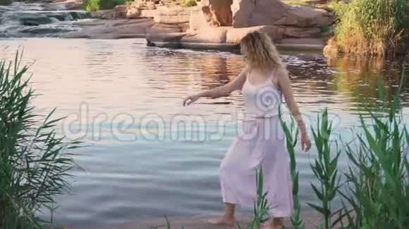 积极的金发女孩赤脚在河边的石头上跳舞就像一条美丽的美人鱼她的粉红色的长而温柔的嬉皮士视频的预览图