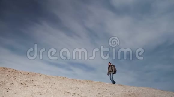 一位戴着眼镜的游客带着一个大背包在美丽的蓝天背景下沿着一个沙丘散步视频的预览图