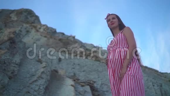 年轻的怀孕女孩穿着条纹红白连衣裙一个女孩站在岩石山脚下抚摸着她的肚子视频的预览图