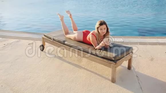 穿着红色比基尼的时尚性感女人躺在甲板椅上使用智能手机应用程序浏览互联网发短信视频的预览图