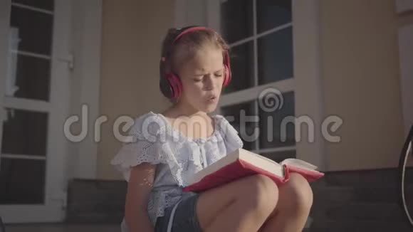 可爱的女孩在暑假里坐在房子附近的院子里听音乐唱歌看书视频的预览图
