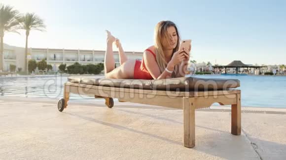 可爱的金发女人穿着红色泳衣在游泳池旁的躺椅上用智能手机浏览互联网视频的预览图