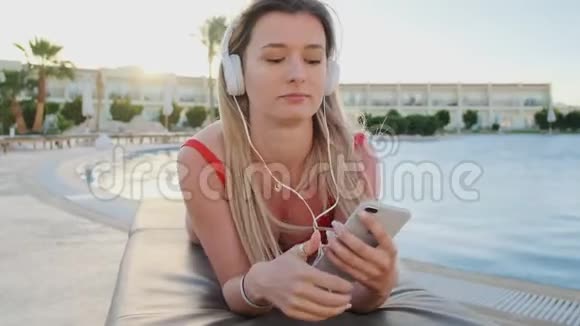 年轻迷人的女士穿着红色比基尼和白色耳机听着智能手机里的音乐一边躺着一边唱歌视频的预览图