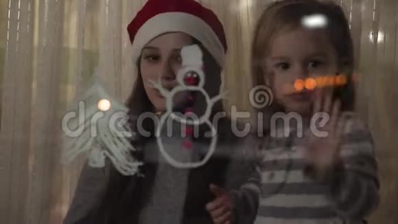 妈妈和女儿在窗玻璃上画一个雪人和一棵圣诞树用丙烯酸在玻璃上画画视频的预览图